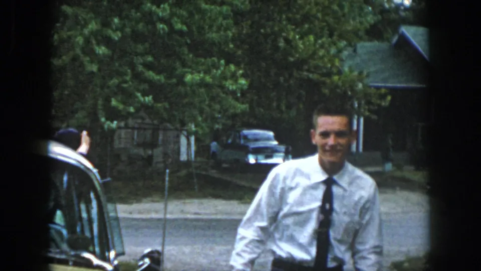A young man in a white shirt and dark tie smiles as he moves away from the car on a leafy residential street in Dayton.