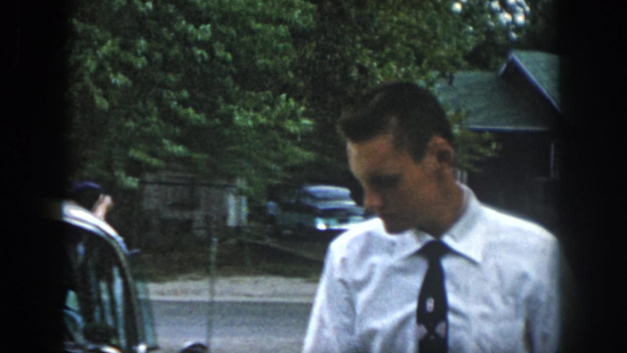 In this 1958 Dayton home-movie frame, a man in church clothes stands near cars outside a leafy residential area.