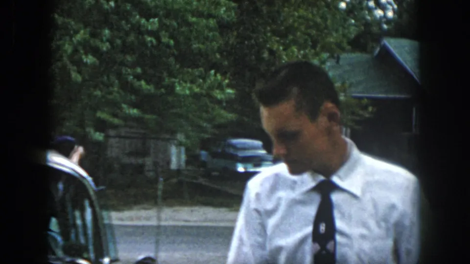 In this 1958 Dayton home-movie frame, a man in church clothes stands near cars outside a leafy residential area.