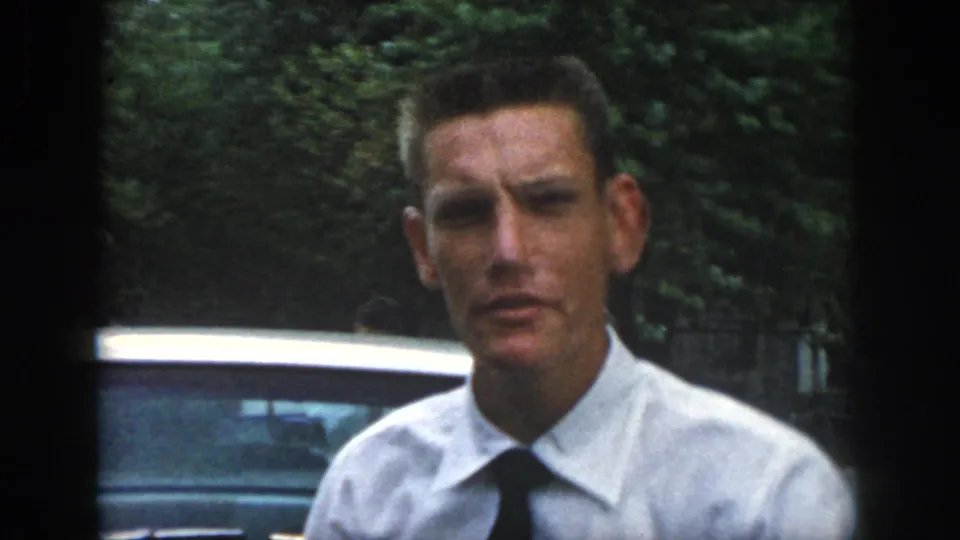 A man in a white dress shirt and dark tie faces the camera near a car in the Dayton, Ohio home-movie scene.