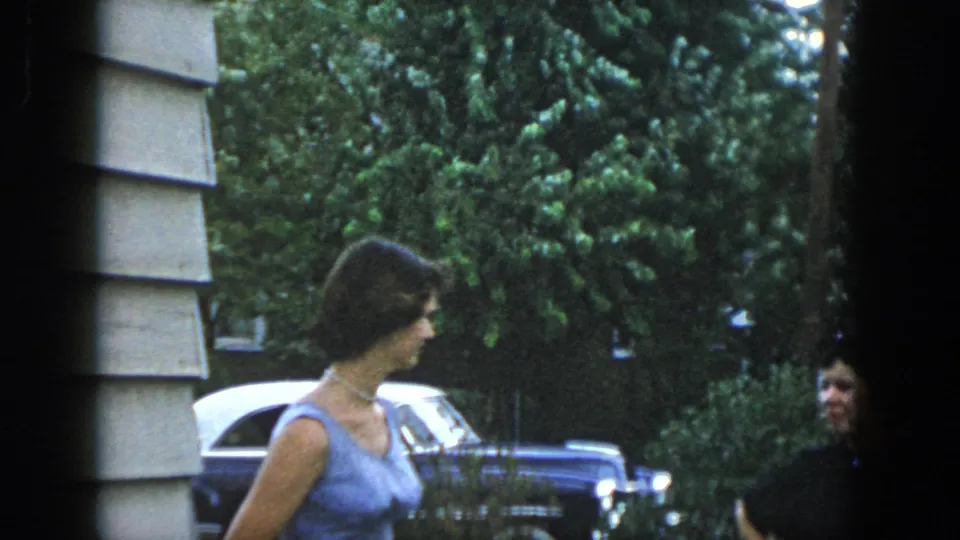 A woman in a blue sleeveless dress stands outside beside cars and another woman in dark clothing in the 1958 Dayton footage.
