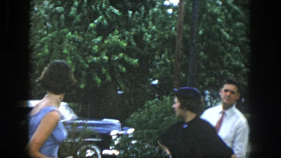 Two women and a man in church clothes gather near mid-century cars in the Dayton residential home-movie scene.