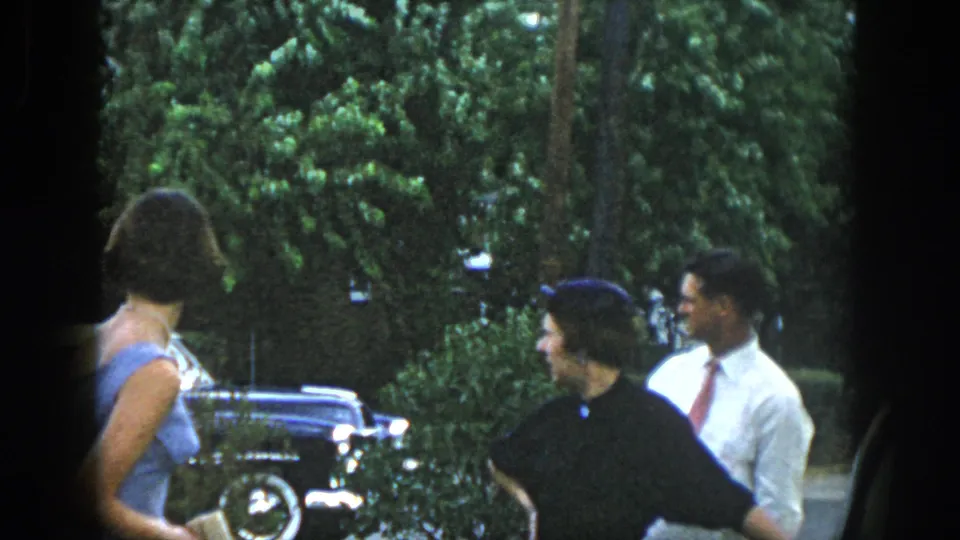 Dayton, Ohio 1958 home-movie frame showing adults in dress clothes talking near a parked classic car and leafy yard.