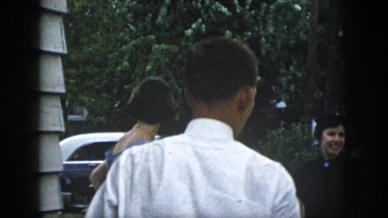 Dayton, Ohio 1958 frame with a man seen from behind while women in formal dresses stand near a parked mid-century car.
