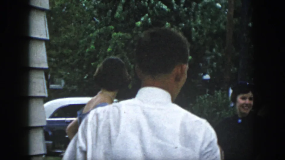 Dayton, Ohio 1958 frame with a man seen from behind while women in formal dresses stand near a parked mid-century car.
