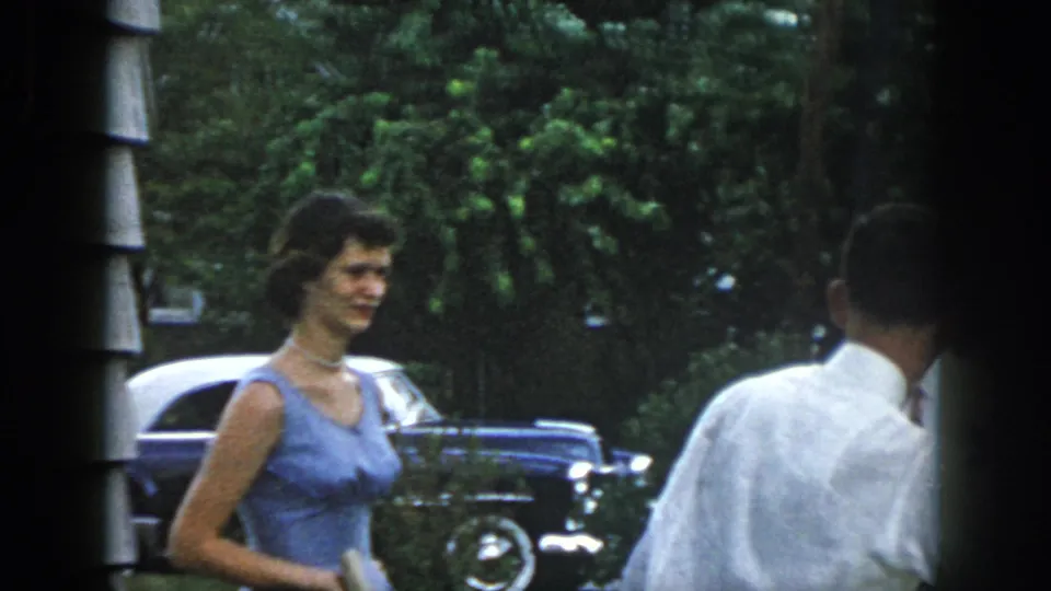 Dayton, Ohio 1958 home-movie frame of a woman in a light blue sleeveless dress near a man in a white shirt and classic cars.