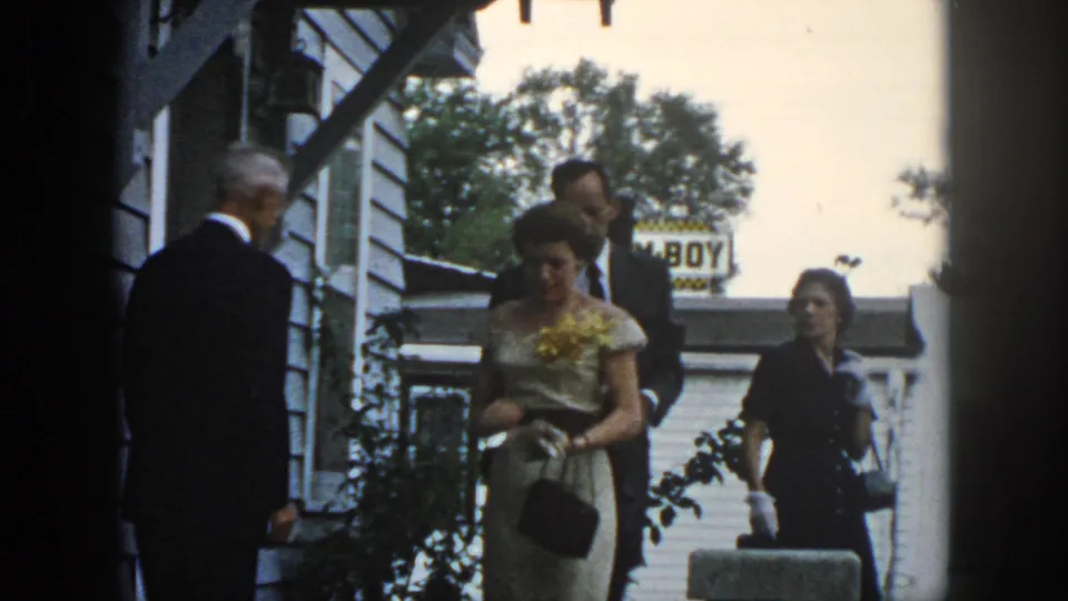 1958 Dayton home-movie frame showing formally dressed adults near a clapboard house, including a woman with a yellow corsage.