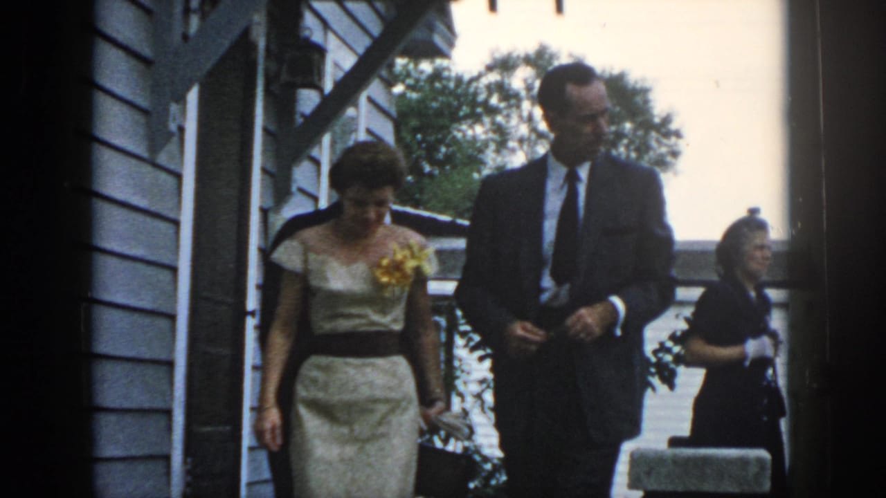 1958 Dayton frame of a dressed couple descending exterior steps beside clapboard siding, with another woman nearby.