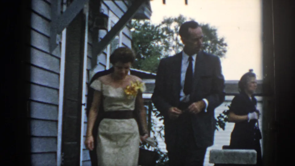 1958 Dayton frame of a dressed couple descending exterior steps beside clapboard siding, with another woman nearby.