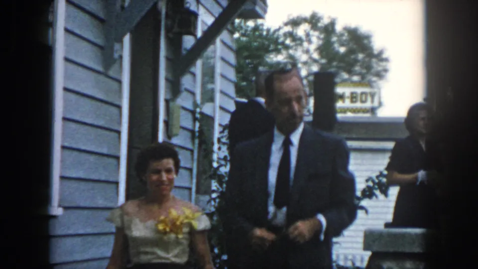 1958 Dayton home-movie frame with a formally dressed couple closer in view as they walk past a clapboard house.