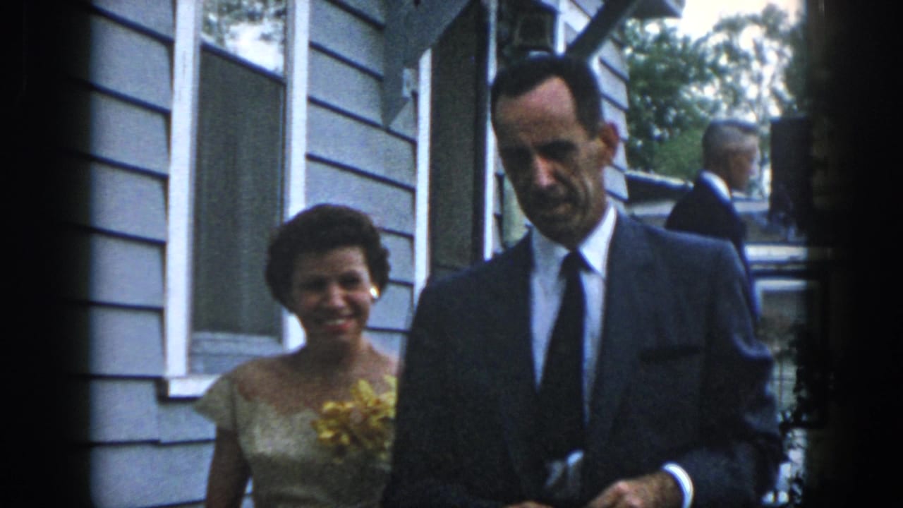 In 1958 Dayton, a woman in a light dress with a yellow flower stands beside a suited man outside a clapboard-sided home.