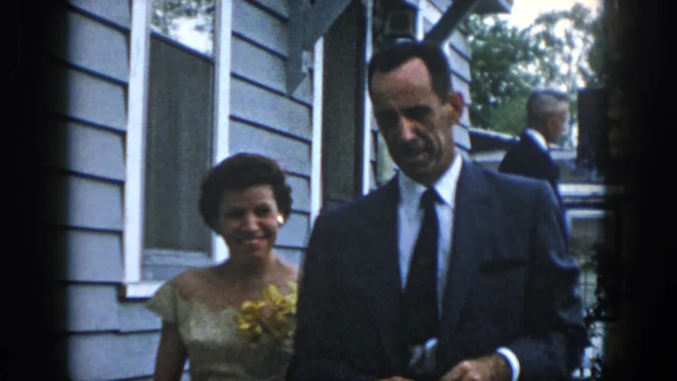 In 1958 Dayton, a woman in a light dress with a yellow flower stands beside a suited man outside a clapboard-sided home.