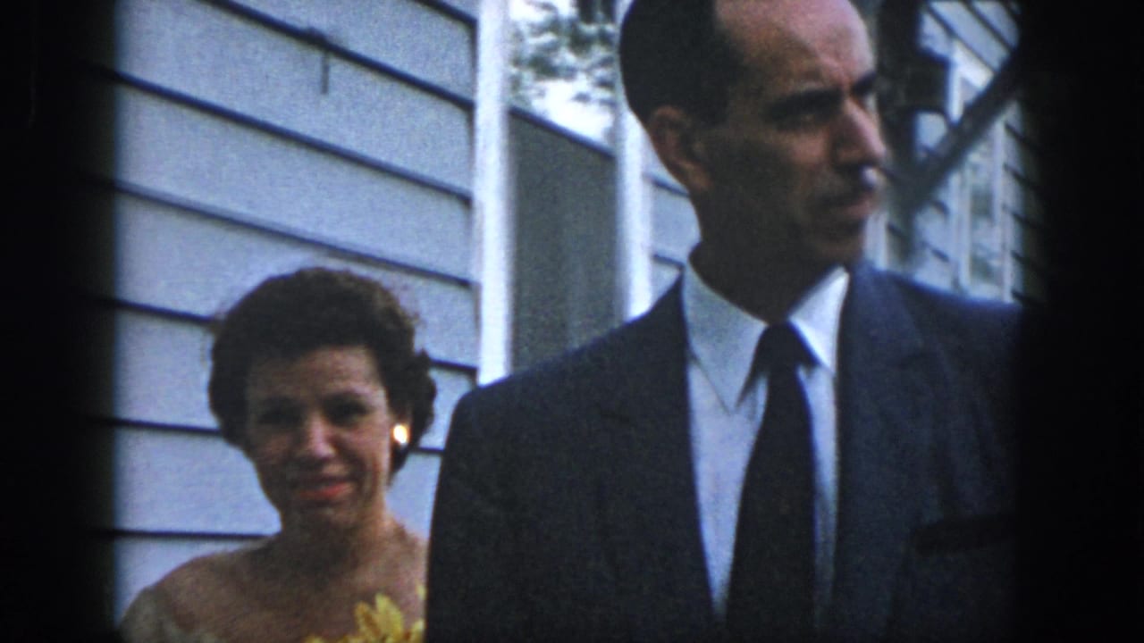 The woman with the yellow flower and the suited man remain near the Dayton house, partly cropped by the handheld 8mm framing.
