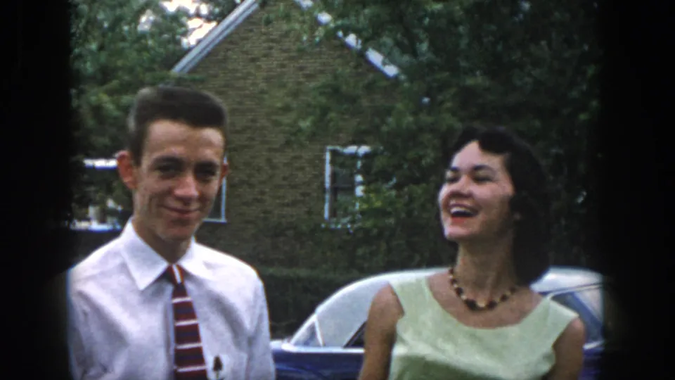 The young couple shares a relaxed smile beside the parked car, with trees and a neighboring house behind them in 1958 Dayton.