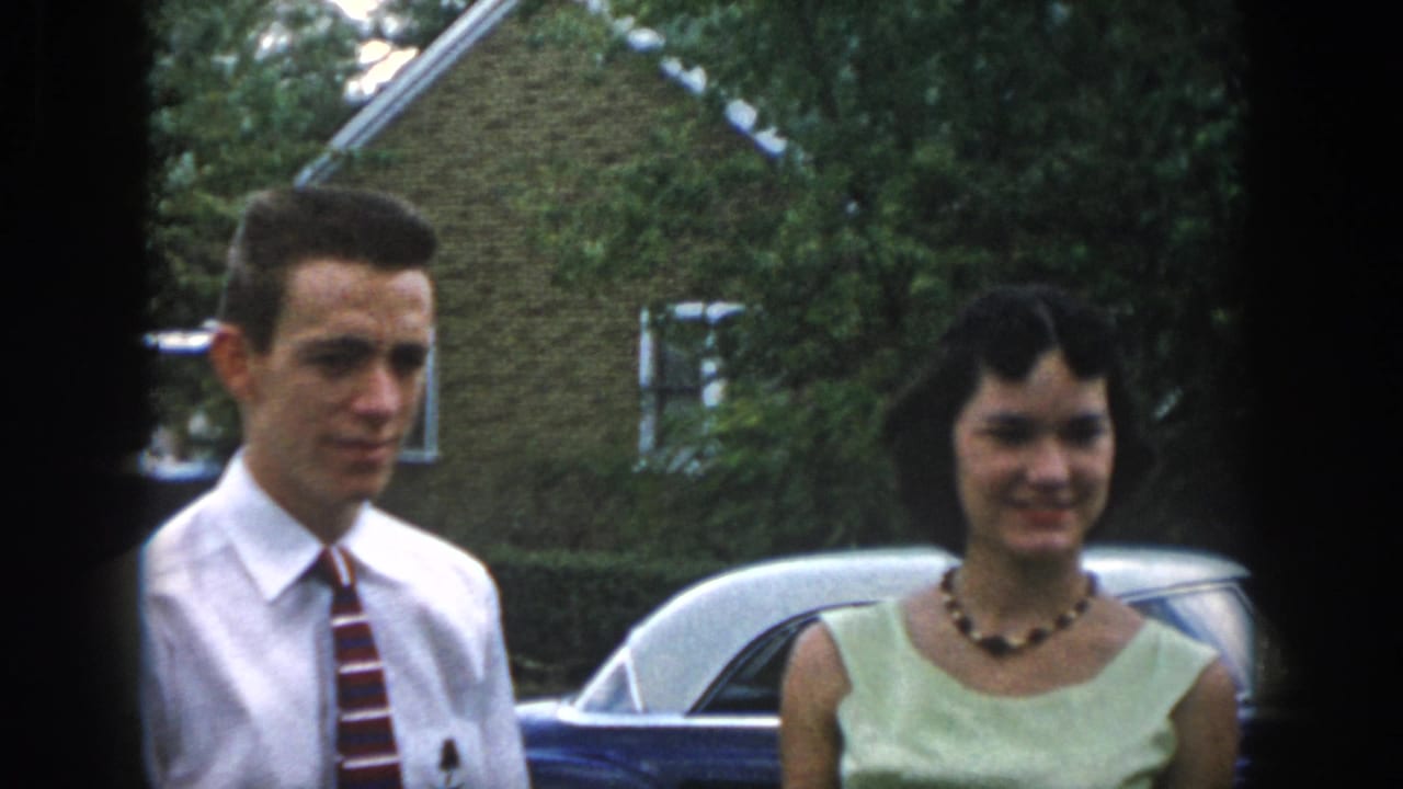 Man in shirt and tie stands beside woman in a light dress near a parked car.
