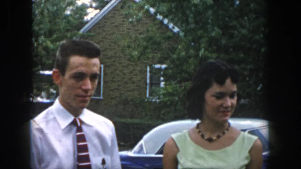 A 1958 Dayton home-movie frame shows the couple in semi-formal clothing beside a car, with the woman glancing down.