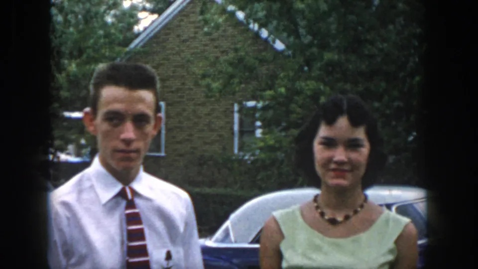 The couple faces the camera in 1958 Dayton, standing before a rounded blue car and brick suburban house.