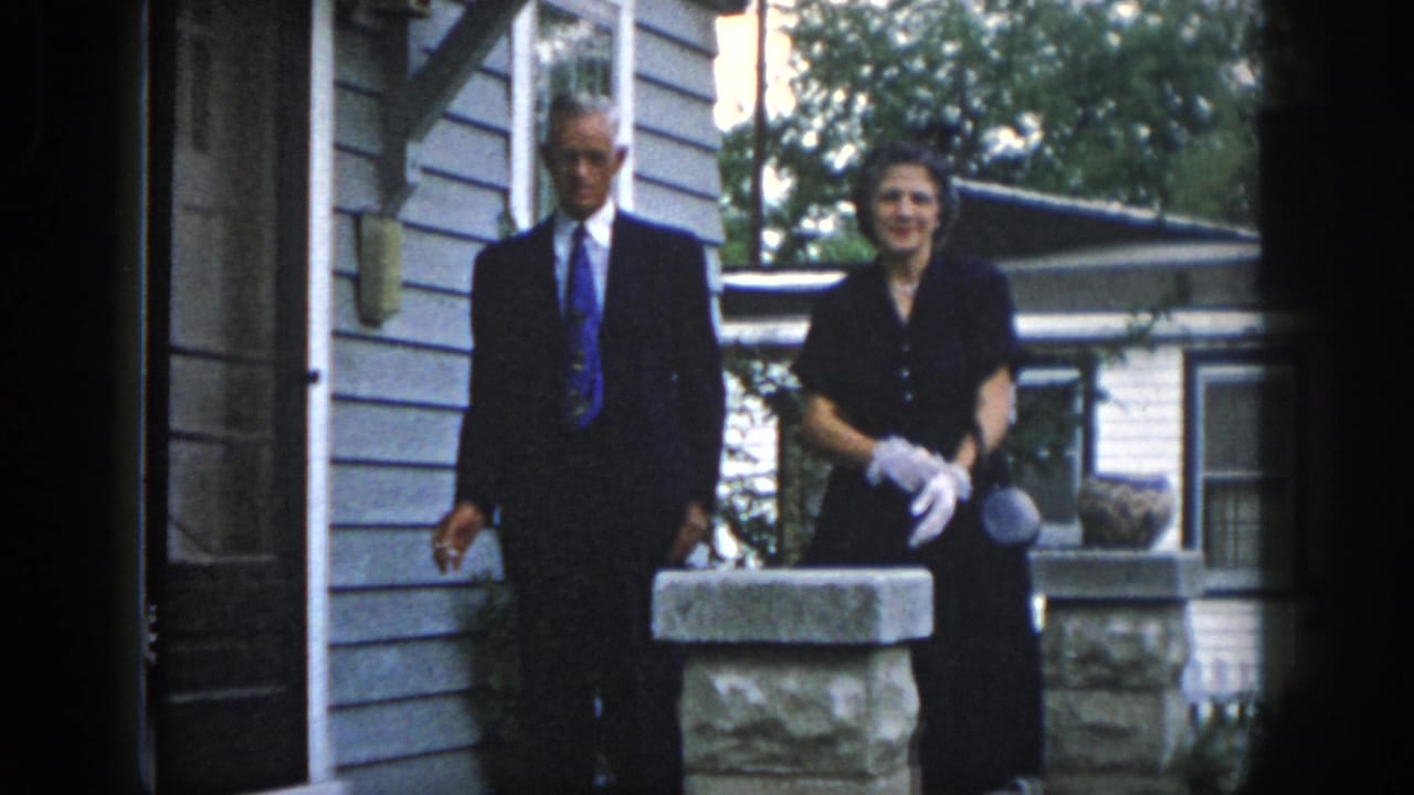 In 1958 Dayton home-movie footage, a man in a dark suit and a woman in a black dress with white gloves stand near a porch pillar.