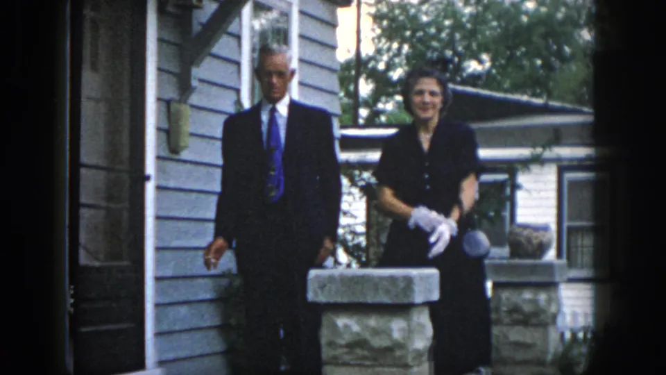 In 1958 Dayton home-movie footage, a man in a dark suit and a woman in a black dress with white gloves stand near a porch pillar.