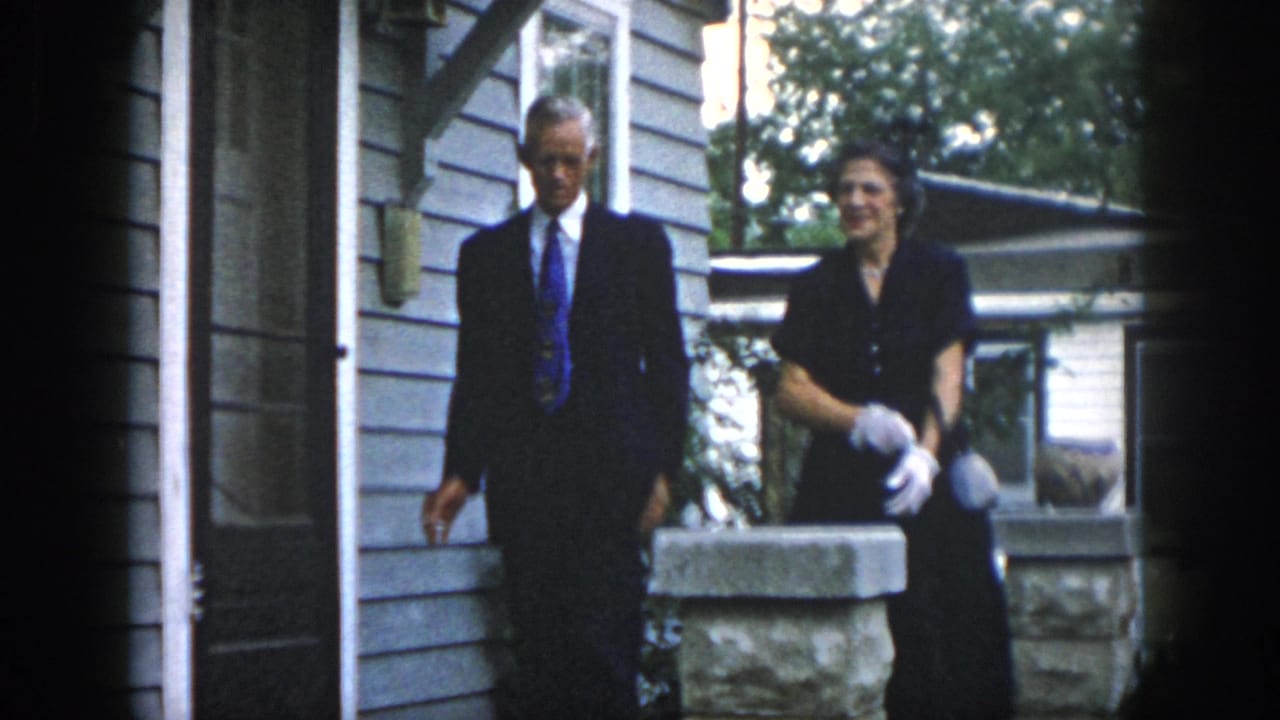Man in a suit walks beside a woman in white gloves near the house exterior.