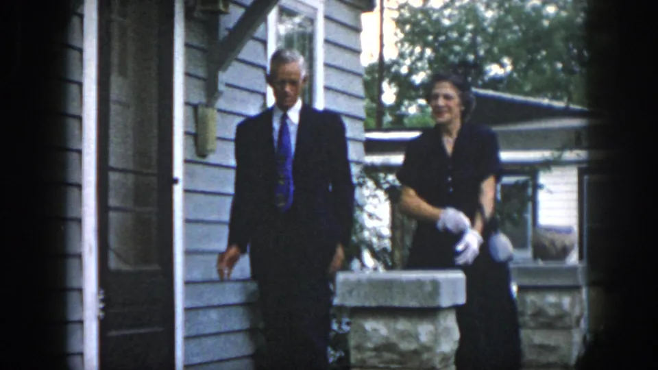 The formally dressed couple moves along the porch area of a Dayton home, with siding, windows, stonework, and trees visible behind them.