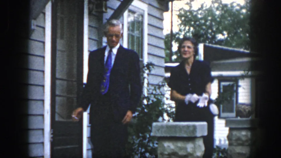 A man in a suit and a woman wearing white gloves stand near the doorway and stone pillar in 1958 Dayton home-movie footage.