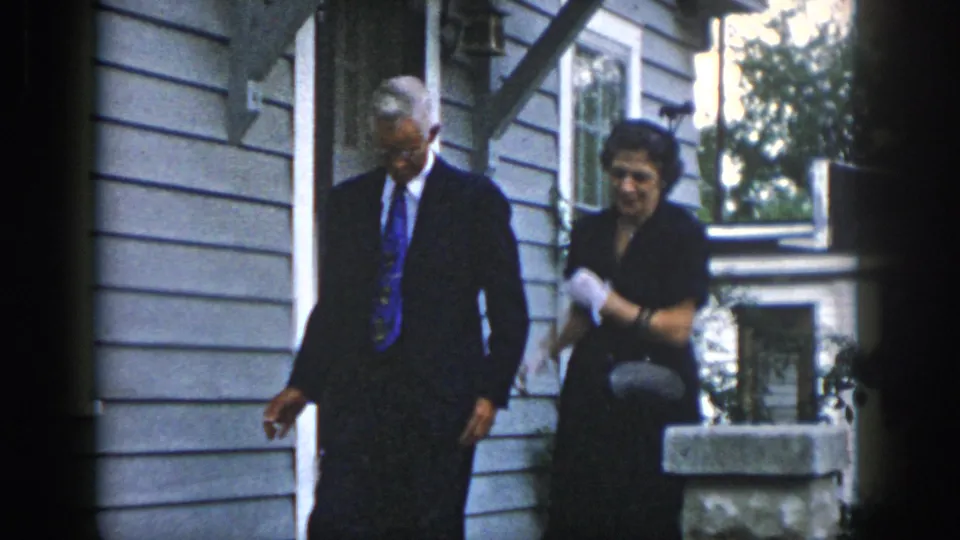 The couple passes the porch pillar, with the woman holding gloves and a purse and the man looking down as they leave the house exterior.