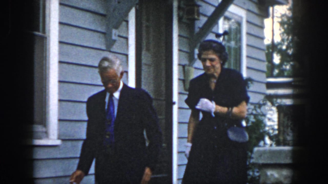 In 1958 Dayton, Ohio home-movie footage, a man in a dark suit and a woman in a dark dress move past a gray clapboard house.