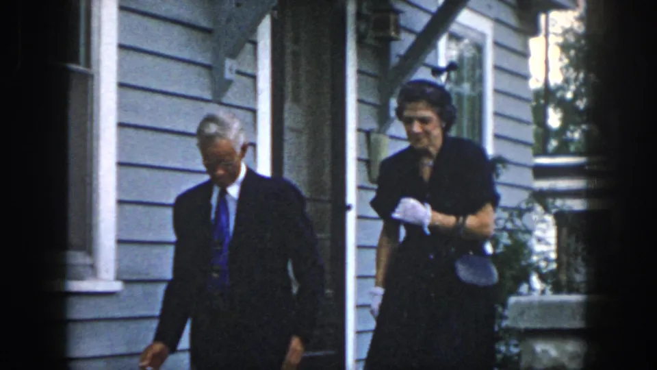 In 1958 Dayton, Ohio home-movie footage, a man in a dark suit and a woman in a dark dress move past a gray clapboard house.