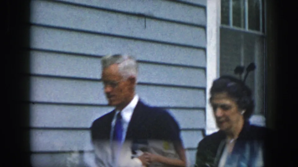 A soft, close 8mm frame shows the suited man and gloved woman moving past the gray house exterior.