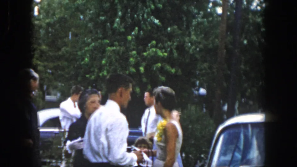 A group gathers outdoors near parked cars and trees, including a man in a white shirt and a woman in a white dress with a yellow lei.