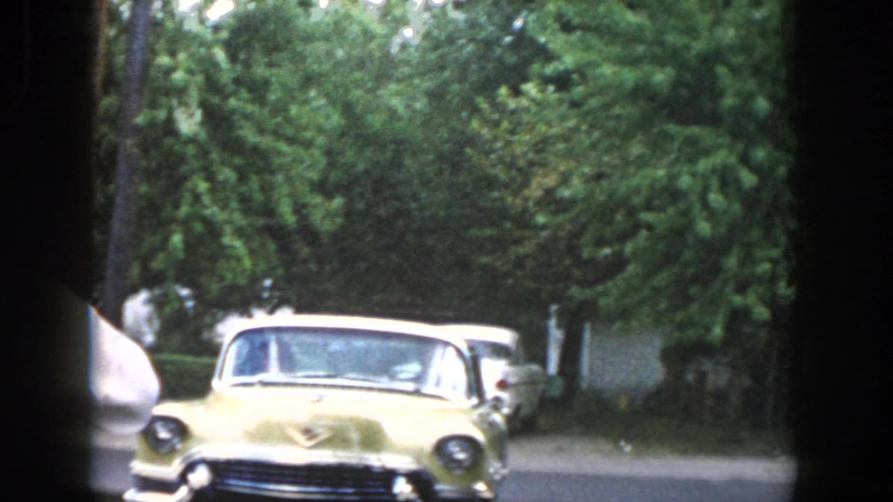 Dayton, Ohio 1958 home-movie frame showing a pale yellow 1950s car near adults at the edge of a tree-lined street.