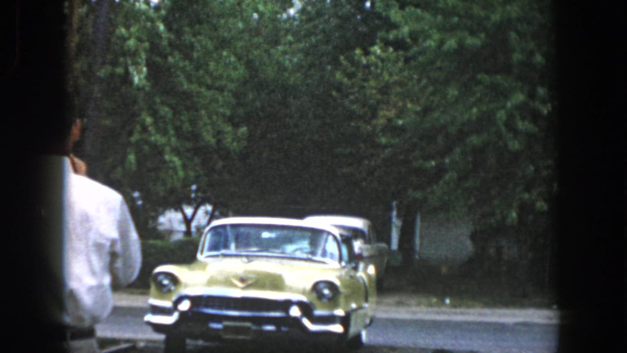 Dayton, Ohio 1958 frame with a yellow 1950s car parked or moving slowly near a man in a light shirt on a leafy street.