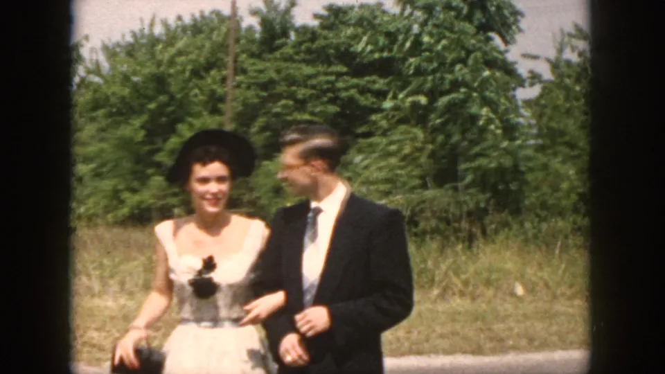 Dayton, Ohio 1958 frame showing a woman in a light dress and dark hat walking with a man in a dark suit outdoors.