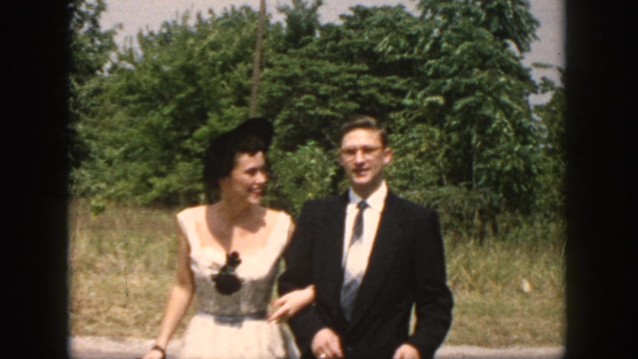 In 1958 Dayton, Ohio home-movie footage, a woman in a pale dress and black hat walks beside a suited man in glasses outdoors.
