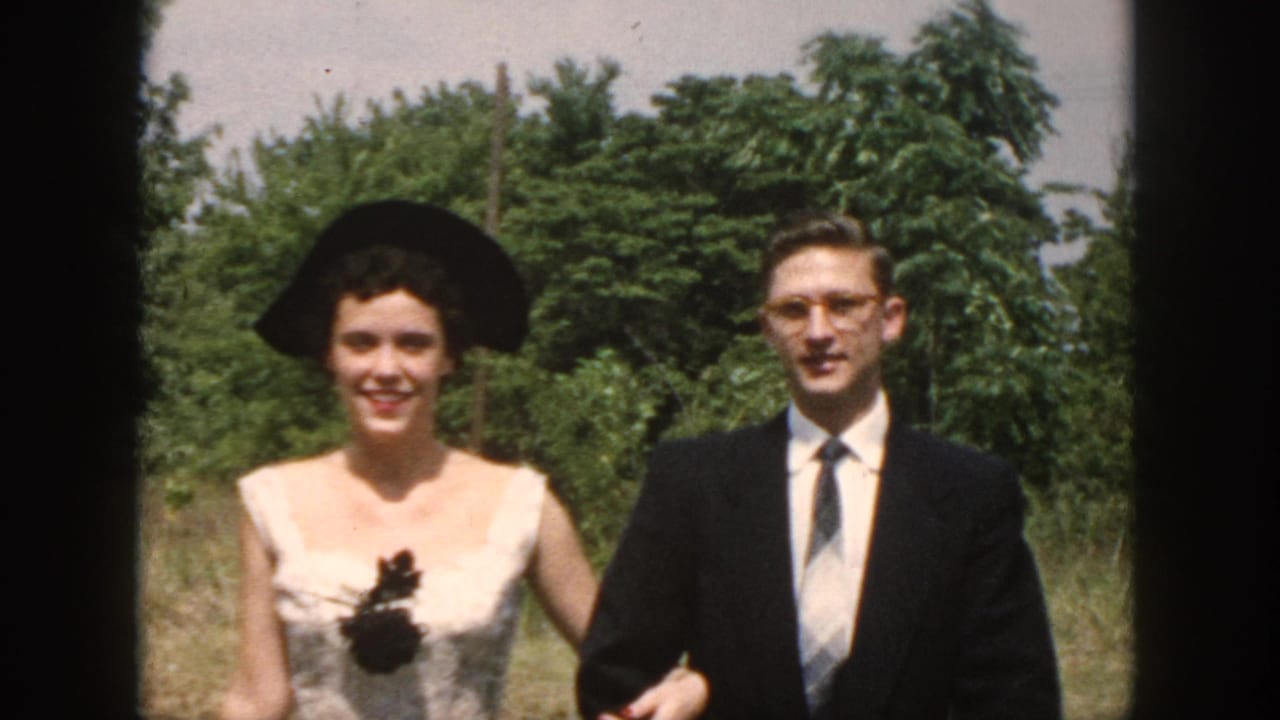 Smiling woman in a wide black hat walks beside a suited man outdoors.
