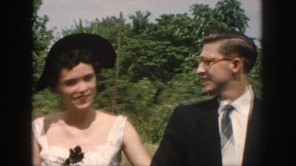 A closer 1958 home-movie frame shows the woman in a pale dress and black hat as the suited man turns his face toward her.