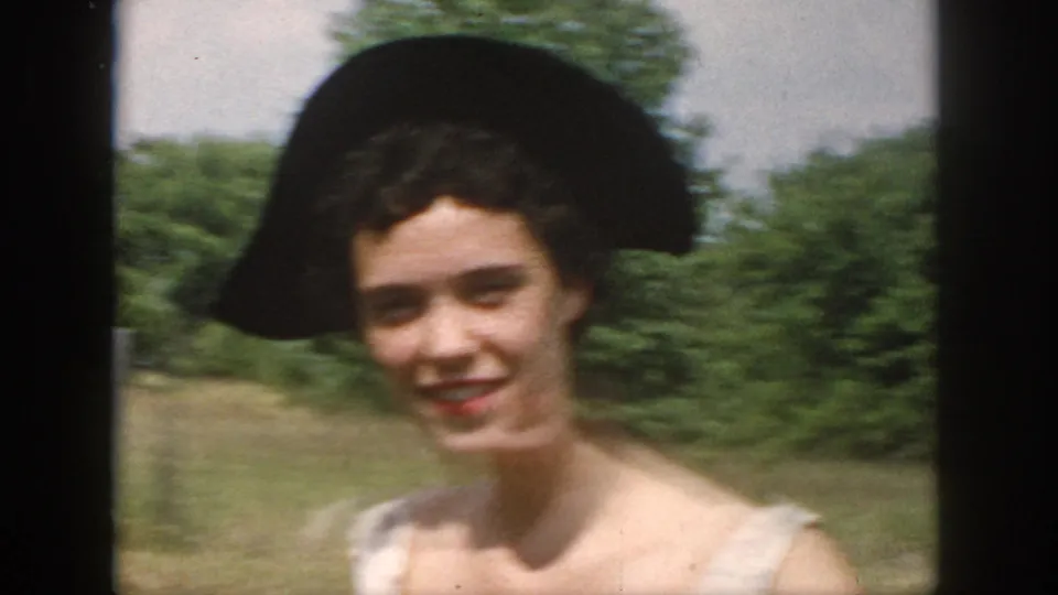 A close 1958 Dayton home-movie frame centers on the smiling woman in a wide black hat and pale dress against a green outdoor background.