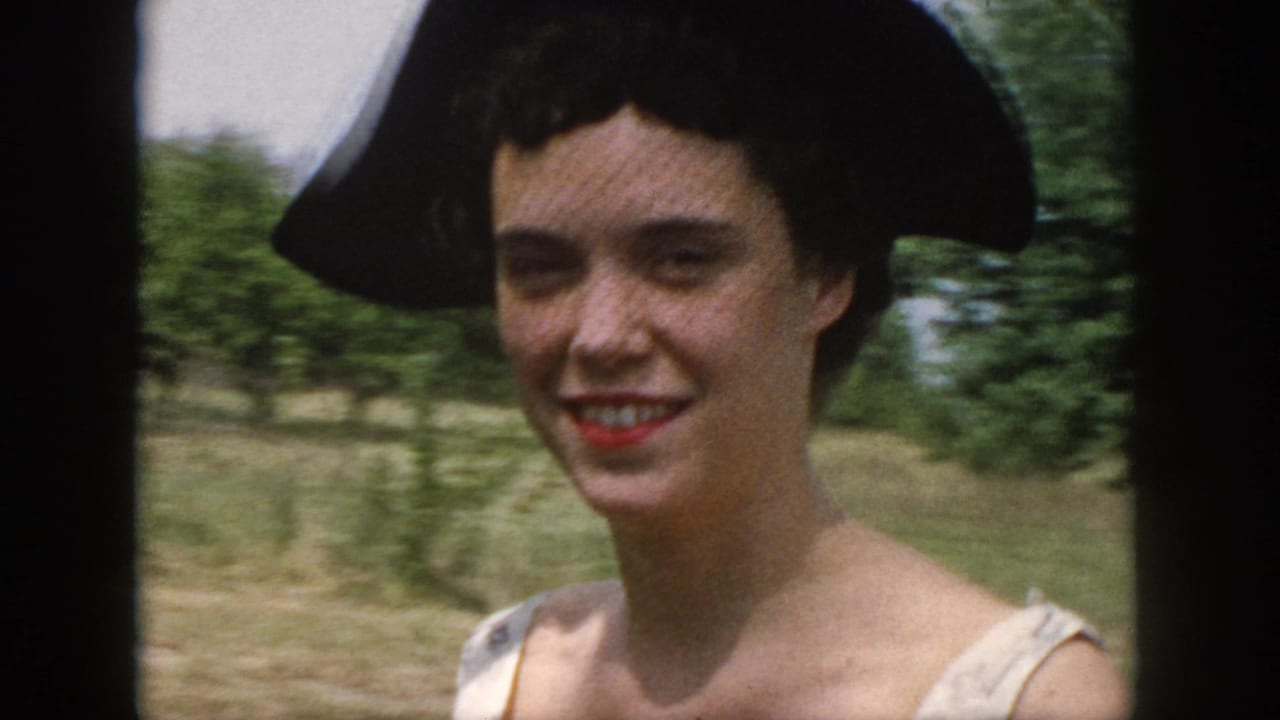 Smiling woman in a black hat and light dress outdoors near trees and grass.