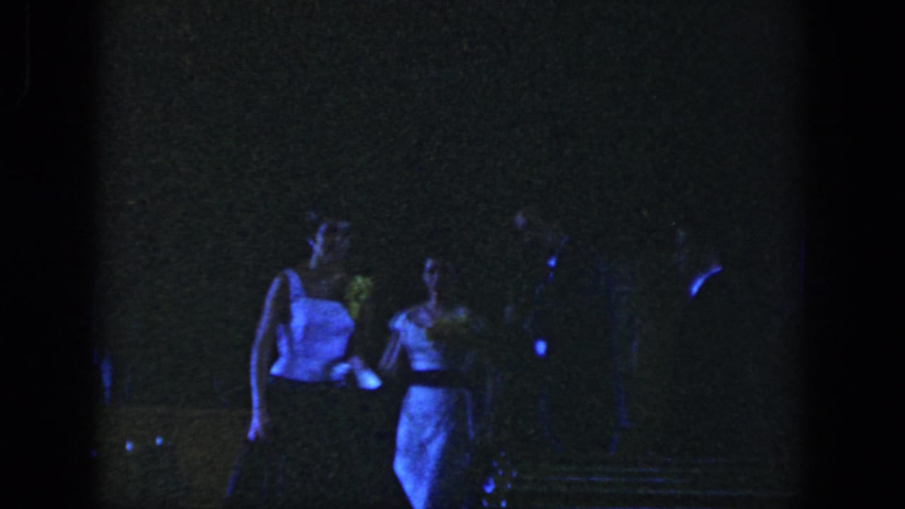 In Dayton 1958 home-movie footage, a young woman in a light formal dress stands among dimly visible guests at night.