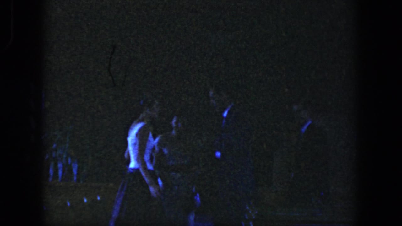 A young man in a light shirt walks through a dark party scene with other figures nearby.