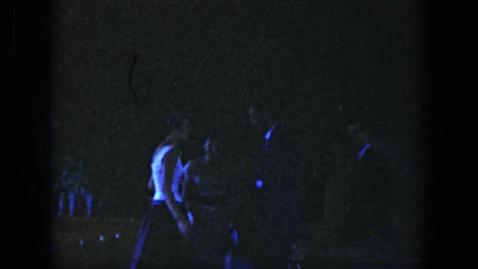 A young man moves through the dim, blue-lit Dayton 1958 party scene as other guests remain faintly visible.