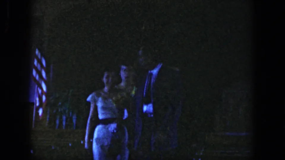 In the 1958 Dayton home-movie frame, a young woman in a light dress stands beside a man amid dim party lighting.
