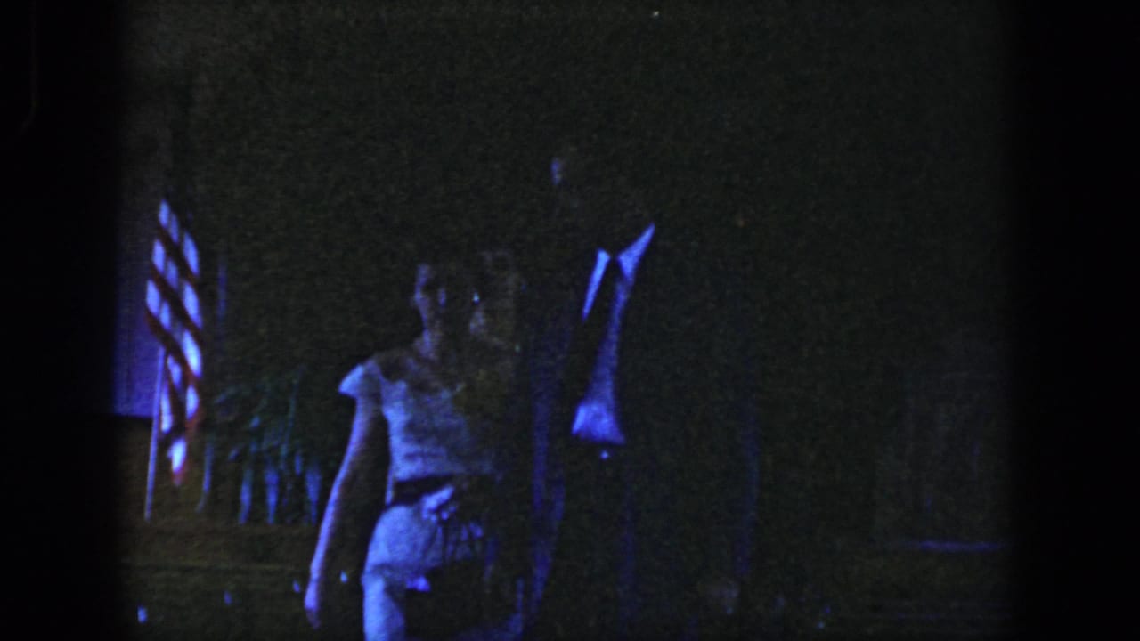 In 1958 Dayton home-movie footage, a suited man and woman in a light dress walk through a dim interior with an American flag behind them.
