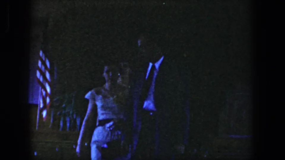 In 1958 Dayton home-movie footage, a suited man and woman in a light dress walk through a dim interior with an American flag behind them.