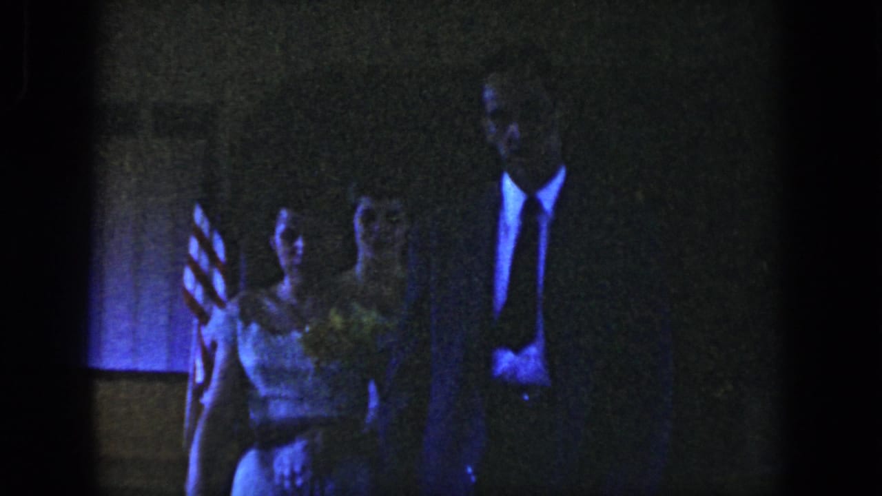 Woman in a light dress and suited man walking toward the camera near an American flag.