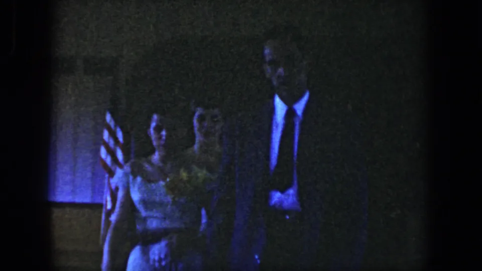 A woman in a light dress and a tall man in a dark suit approach the camera inside a Dayton, Ohio venue with a U.S. flag visible.