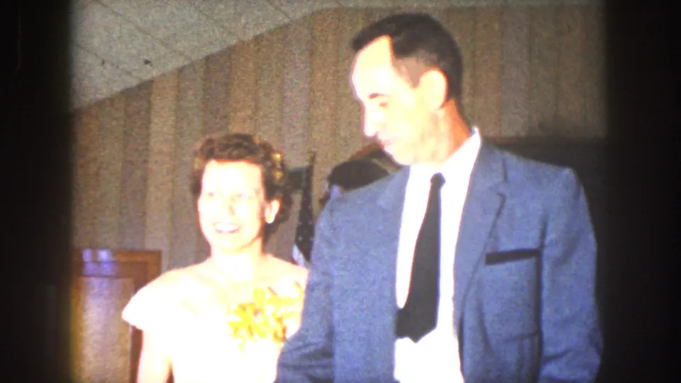The couple is more clearly lit, with the woman smiling in a light dress and the man smiling in a suit inside a wood-paneled room.