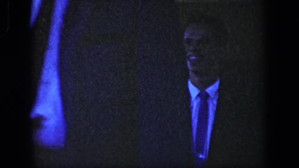 A low-light frame shows the suited man near the camera, with part of the woman's light dress visible at the left edge.