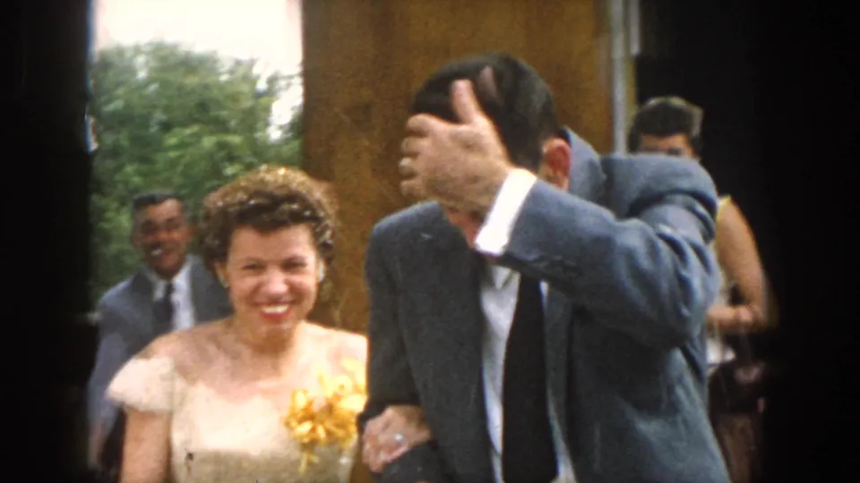 The bride smiles in a light off-the-shoulder dress with a yellow floral accent while the groom shields his head during the wedding exit.